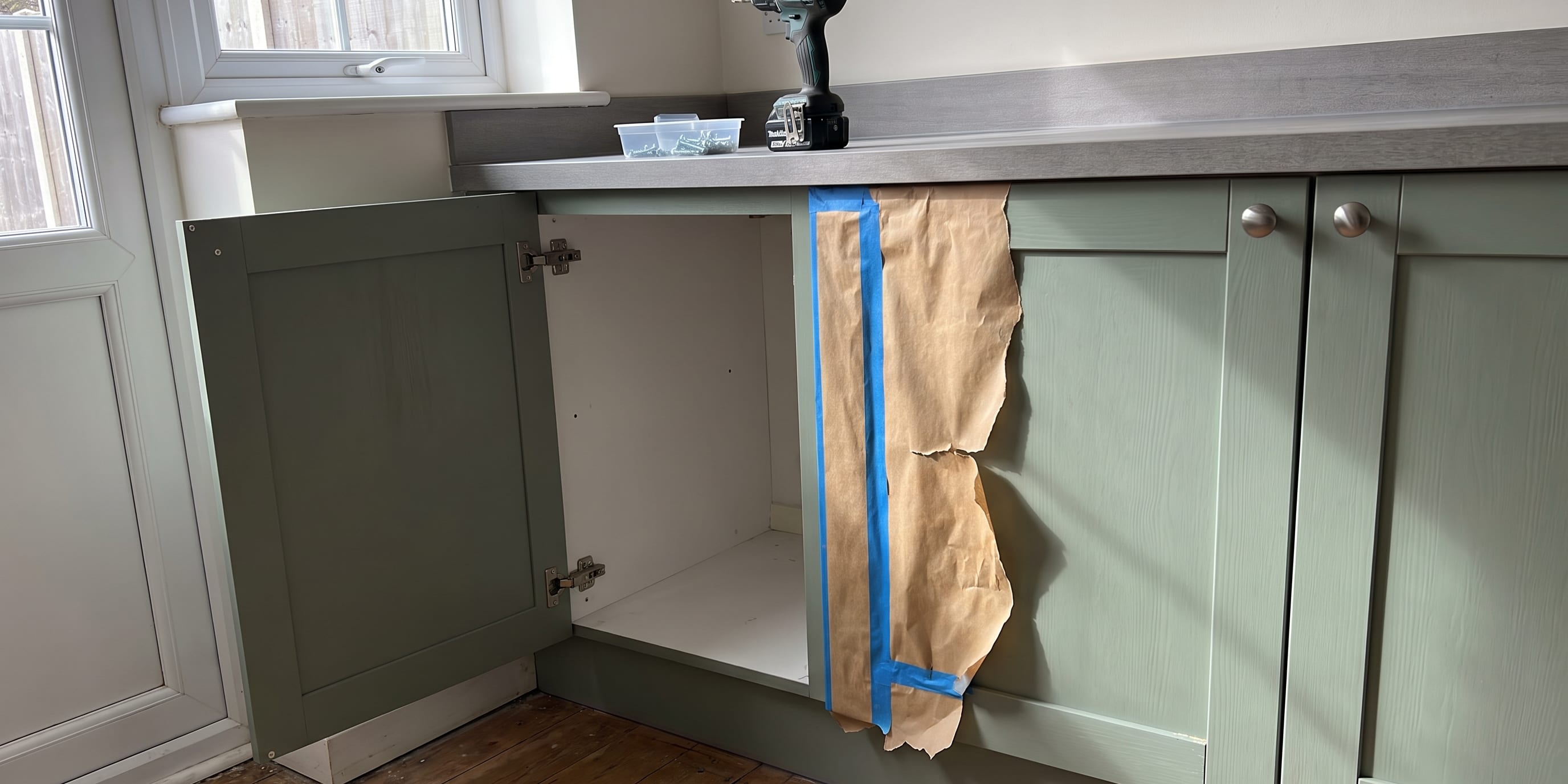 A kitchen cabinet run where one green shaker door has been removed and leans against the counter with its hinges exposed, while the door next to it is still attached but partly covered in blue masking tape and brown paper.