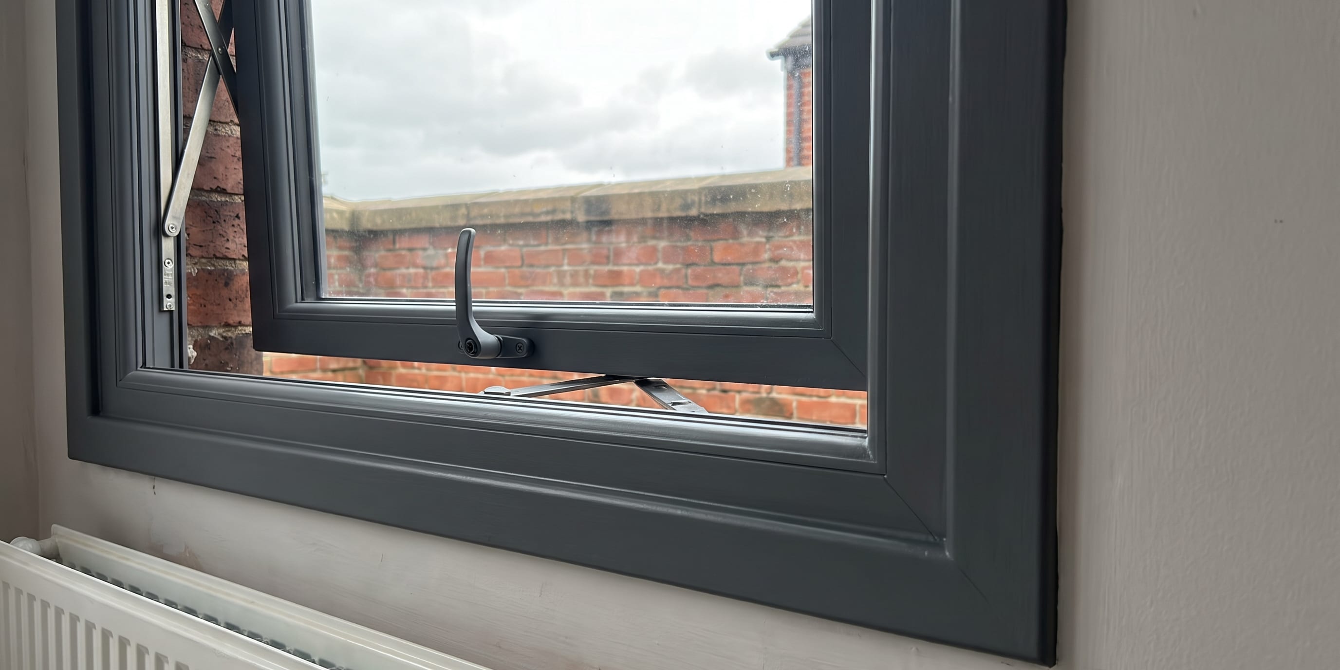 An open grey uPVC casement window with a hinge stay and handle, viewed from inside a room with a white radiator beneath the sill and a brick wall visible outside.
